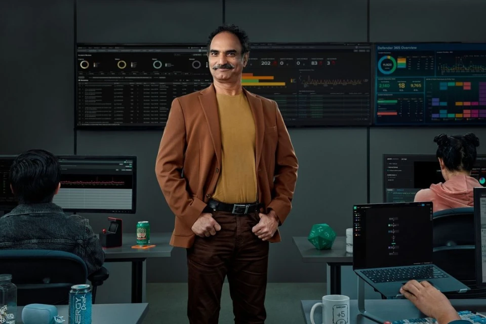Man in brown jacket smiles confidently in a tech office environment surrounded by computer screens and colleagues working at their desks.